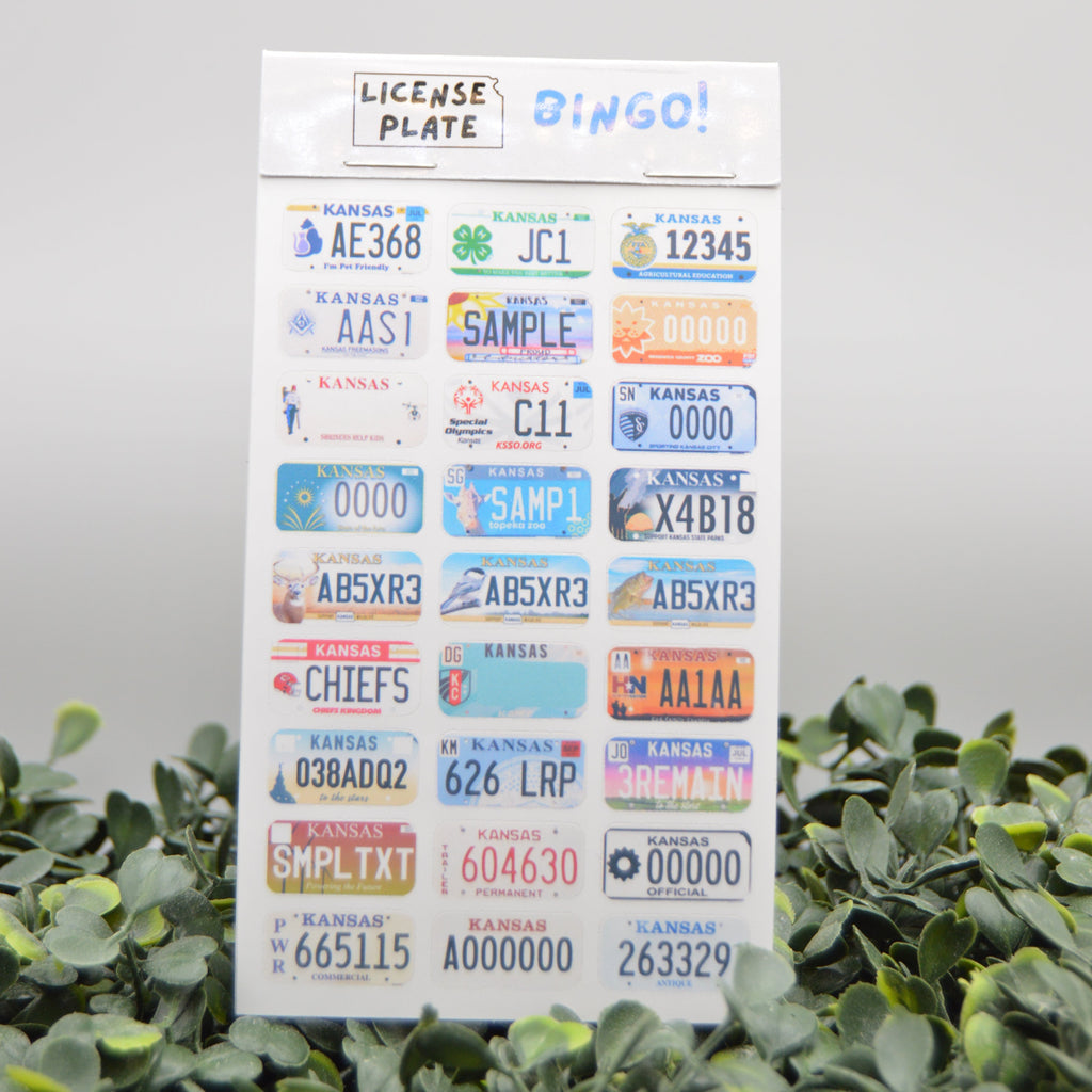 A sheet of novelty license plate stickers displayed upright on green foliage against a light gray background. The sticker sheet has a white header that reads 'LICENSE PLATE' and 'DINGO!' with multiple colorful sample license plates printed below.