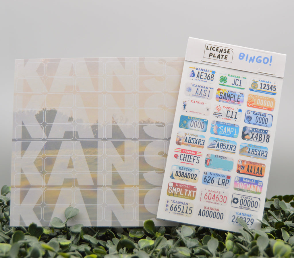 A sheet of decorative license plate stickers labeled 'BINGO!' stands upright next to a clear plastic storage box with a large 'KANSAS' word design on the front, set on green foliage against a soft gray background.