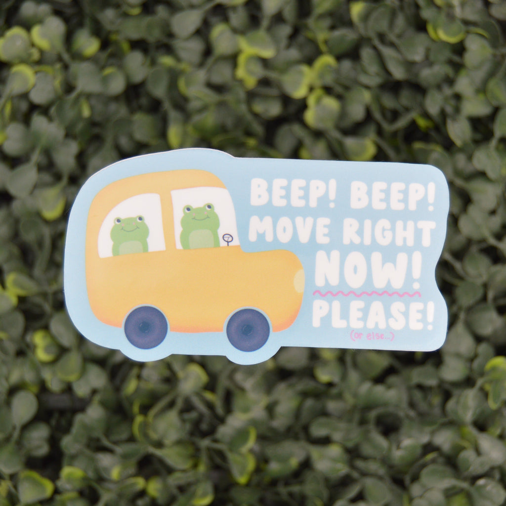 A die-cut decorative sign shaped like a yellow bus with two green cartoon frogs inside, placed against a blurred green hedge background. The sign reads, 'BEEP! BEEP! MOVE RIGHT NOW! PLEASE!'.