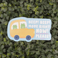 A die-cut decorative sign shaped like a yellow bus with two green cartoon frogs inside, placed against a blurred green hedge background. The sign reads, 'BEEP! BEEP! MOVE RIGHT NOW! PLEASE!'.