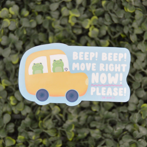 A die-cut decorative sign shaped like a yellow bus with two green cartoon frogs inside, placed against a blurred green hedge background. The sign reads, 'BEEP! BEEP! MOVE RIGHT NOW! PLEASE!'.