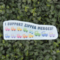 A die-cut sticker with the text 'I SUPPORT ZIPPER MERGES!' and small colorful cartoon cars, shown against a blurred green hedge background.