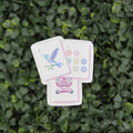 Three decorative cards with colorful illustrations of a hummingbird, flowers, and an owl, arranged on a background of dense green foliage.