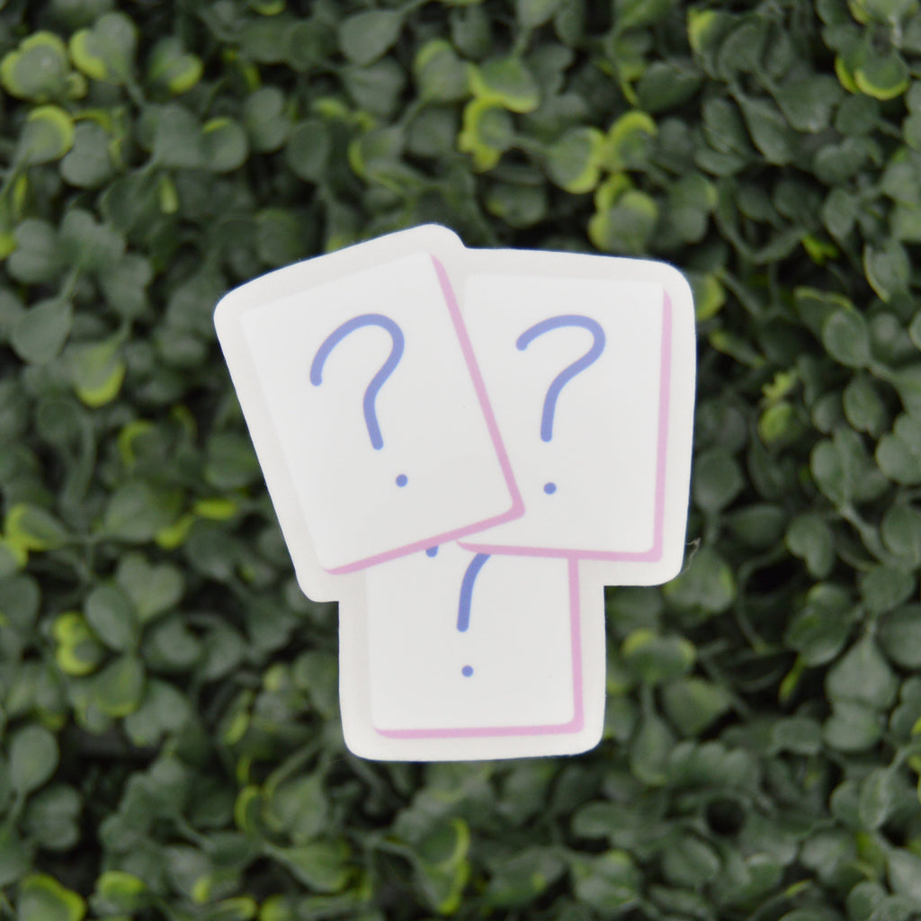 Three white square cards with blue question marks and pink borders, arranged in a small stack against a dense green leafy background.