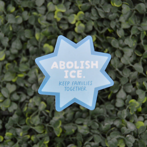 A light blue star-shaped sign with the text 'ABOLISH ICE. KEEP FAMILIES TOGETHER' in white, placed against a dense green leafy hedge background.