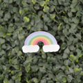 A small rainbow-shaped accessory with white cloud ends, placed against a dense green leafy background.