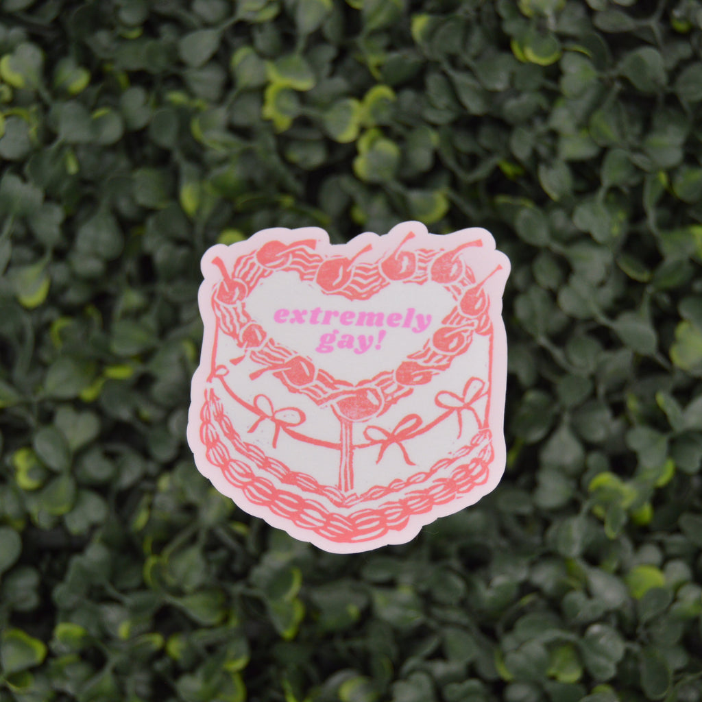 A die-cut sticker featuring a pink line art illustration of a cake with the words 'extremely sad!' in pink text, placed against a background of green leafy foliage.