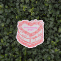 A die-cut sticker featuring a pink line art illustration of a cake with the words 'extremely sad!' in pink text, placed against a background of green leafy foliage.