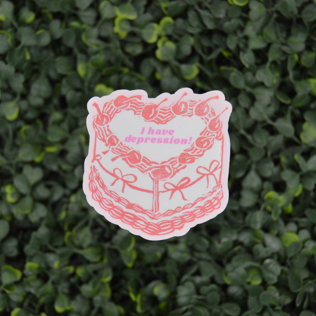 A die-cut sticker featuring a pink and white illustrated cake with the text 'I have depression!' in pink, placed against a background of dense green ivy.