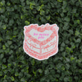 A die-cut sticker featuring a pink and white illustrated cake with the text 'be mine pis' in pink, placed against a background of green leafy foliage.