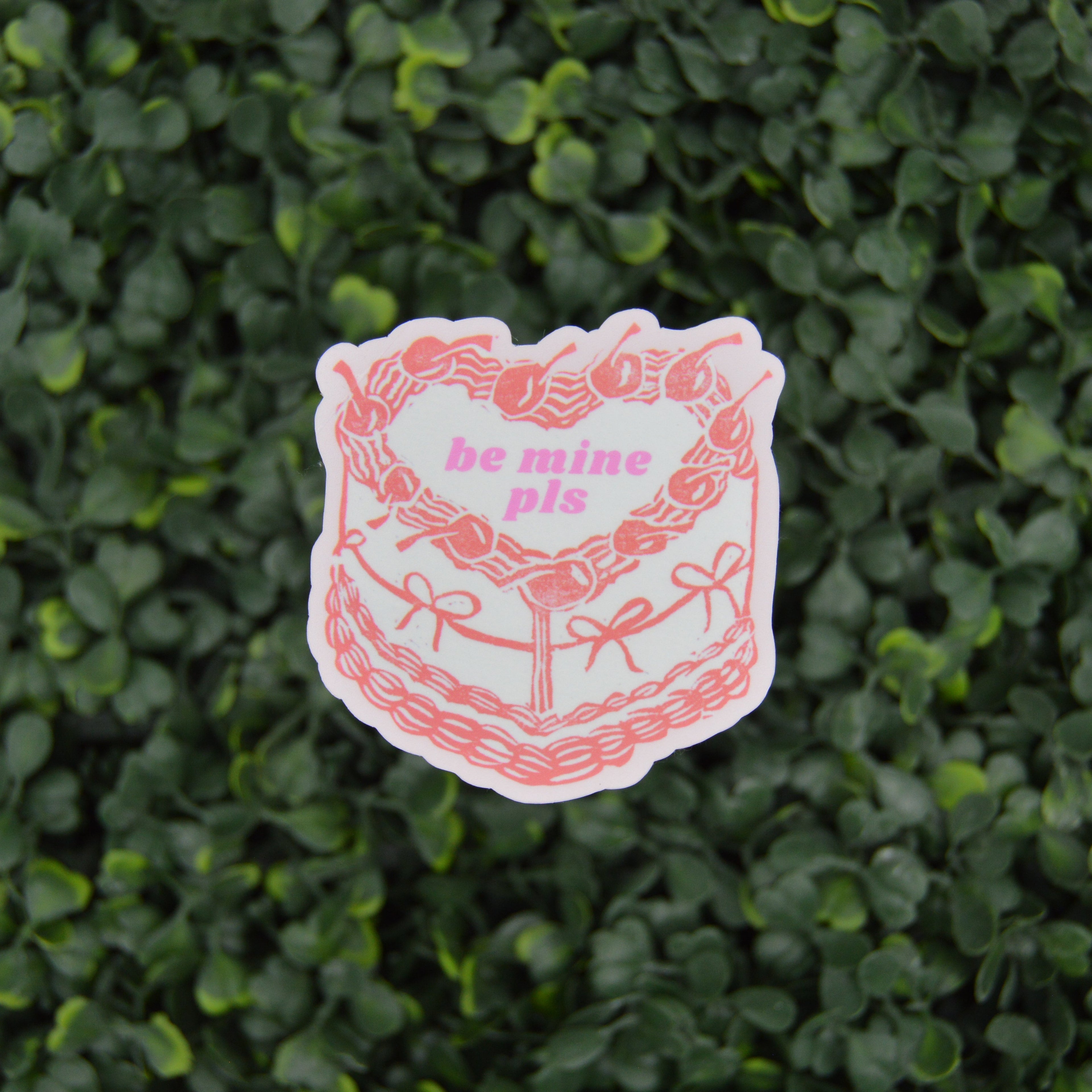 A die-cut sticker featuring a pink and white illustrated cake with the text 'be mine pis' in pink, placed against a background of green leafy foliage.