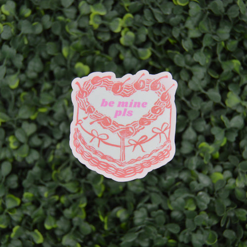 A die-cut sticker featuring a pink and white illustrated cake with the text 'be mine pis' in pink, placed against a background of green leafy foliage.