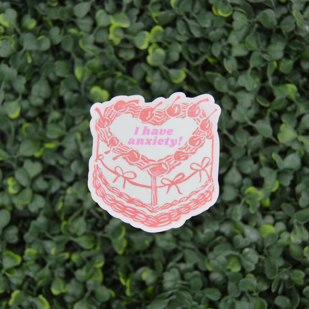 A die-cut sticker featuring a pink and white illustration of a cake with the text 'I have anxiety!' in pink, placed against a background of green foliage.