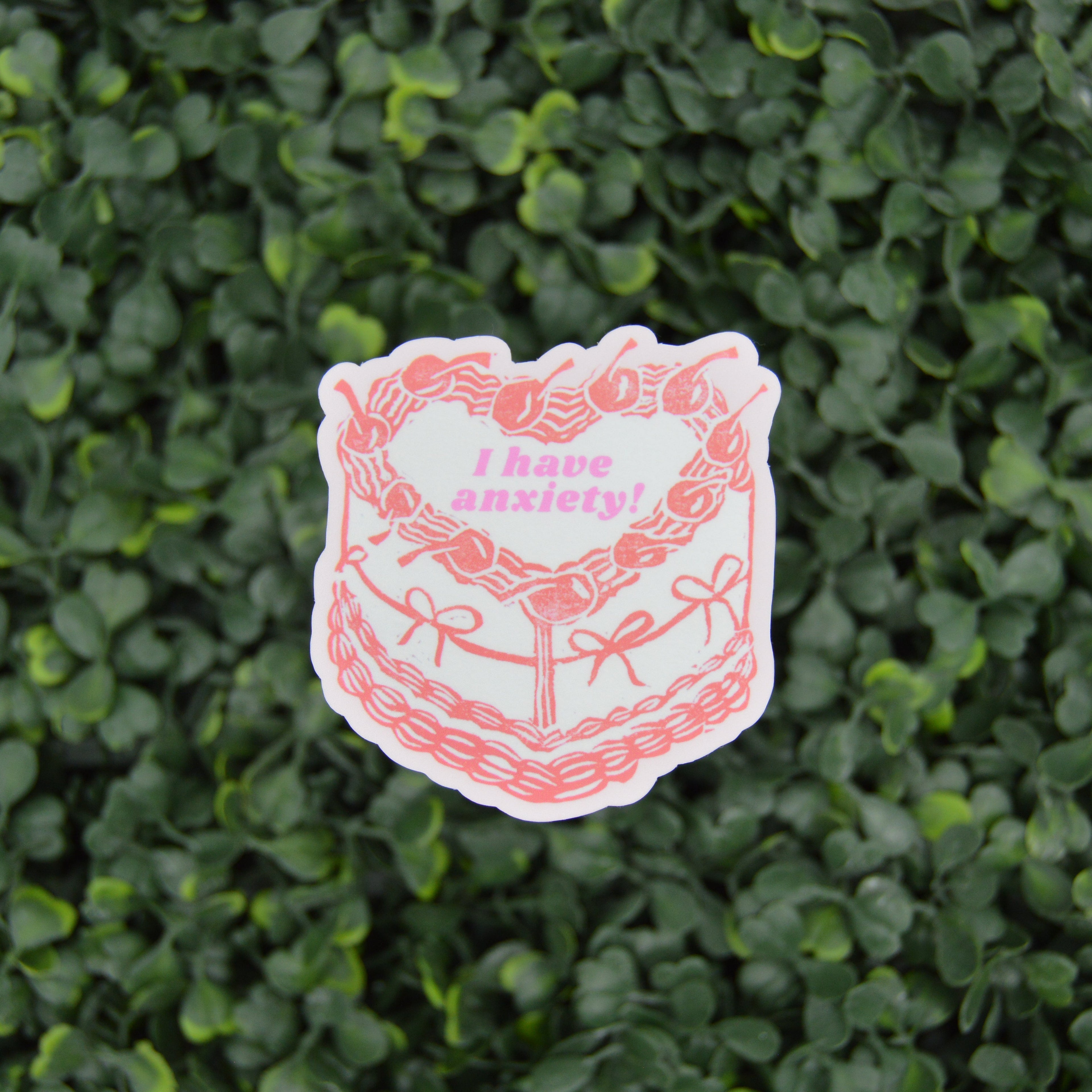 A die-cut sticker featuring a pink and white illustration of a cake with the text 'I have anxiety!' in pink, placed against a background of green foliage.