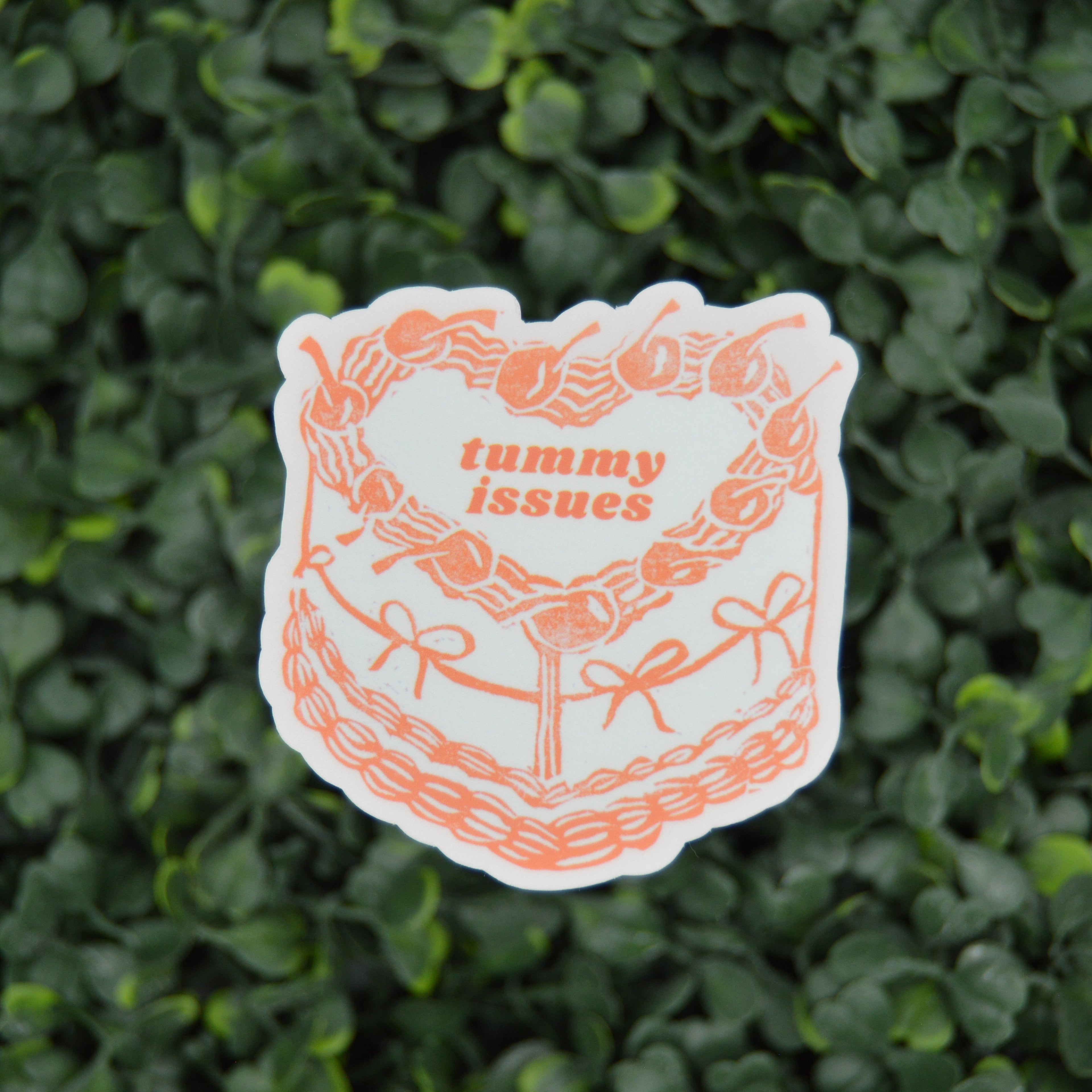 A die-cut sticker featuring an orange line-art illustration of a cake with the words 'tummy issues' in orange text, placed against a background of green foliage.