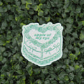 A die-cut sticker featuring a green line art illustration of a cake with the text 'apple of my eye', placed on a textured green leaf wall background.