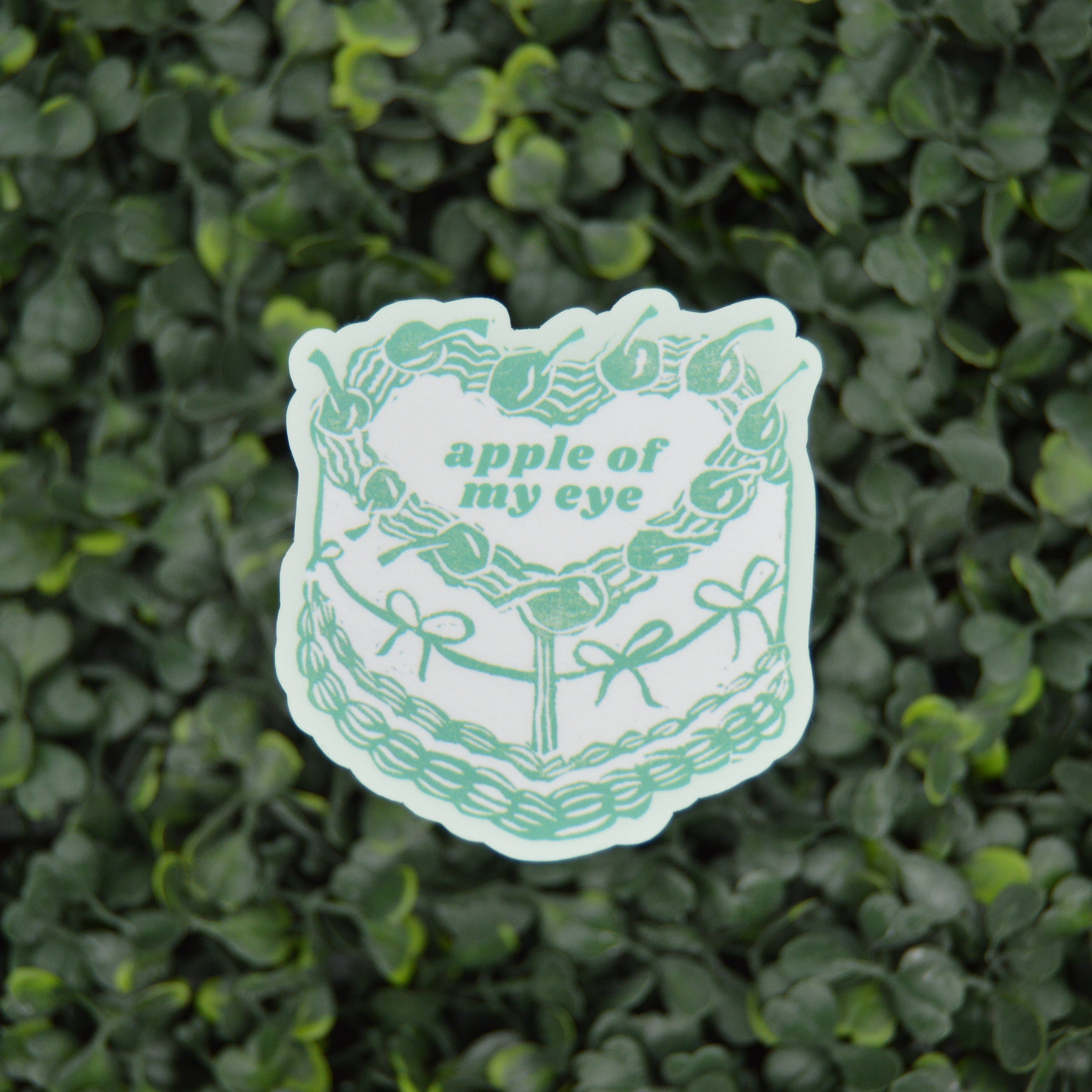 A die-cut sticker featuring a green line art illustration of a cake with the text 'apple of my eye', placed on a textured green leaf wall background.