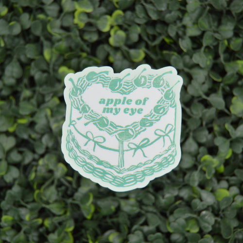 A die-cut sticker featuring a green line art illustration of a cake with the text 'apple of my eye', placed on a textured green leaf wall background.