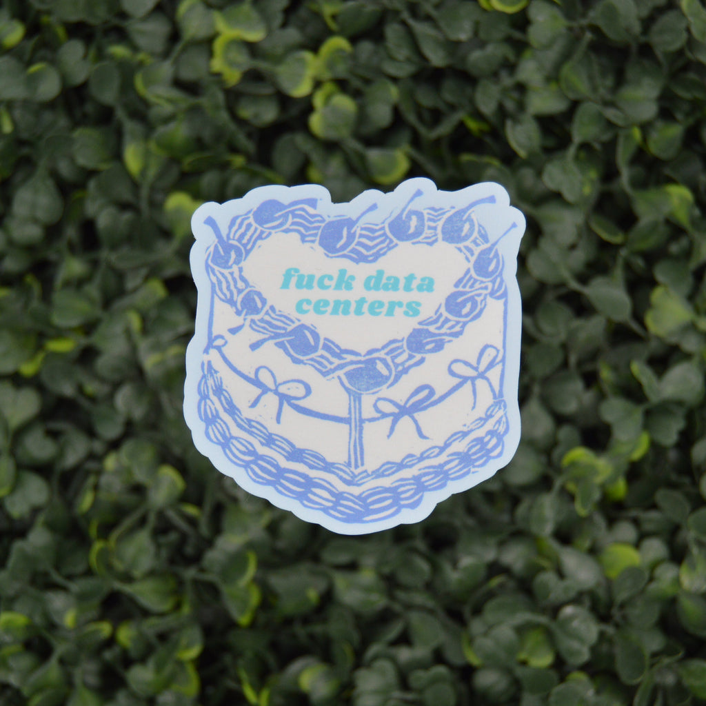 A die-cut sticker featuring a blue and white illustrated cake with the text 'fuck data centers' in light blue, placed against a background of green variegated foliage.