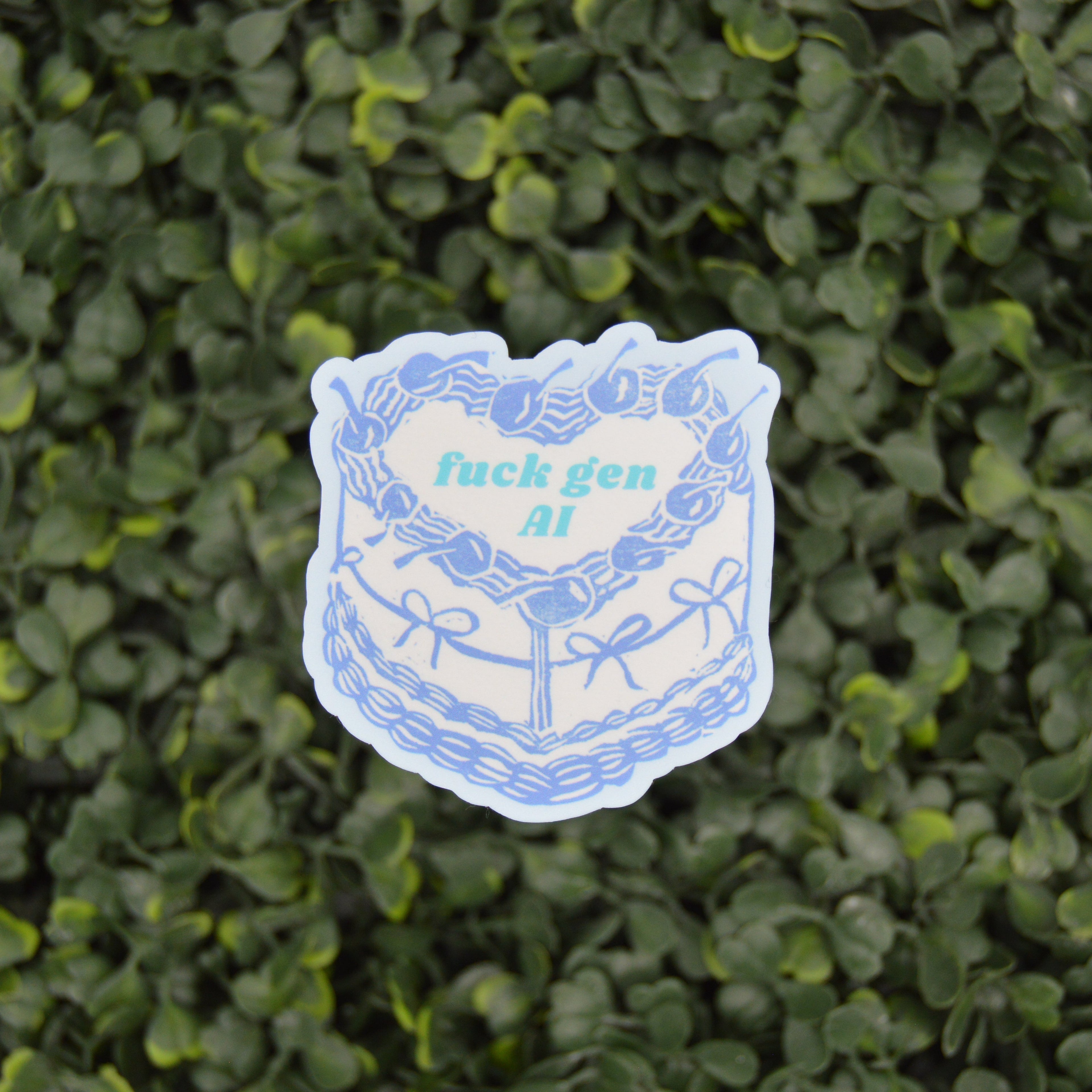 A die-cut sticker featuring a blue and white illustrated cake with the text 'fuck gen AI' in light blue, placed against a background of green ivy leaves.