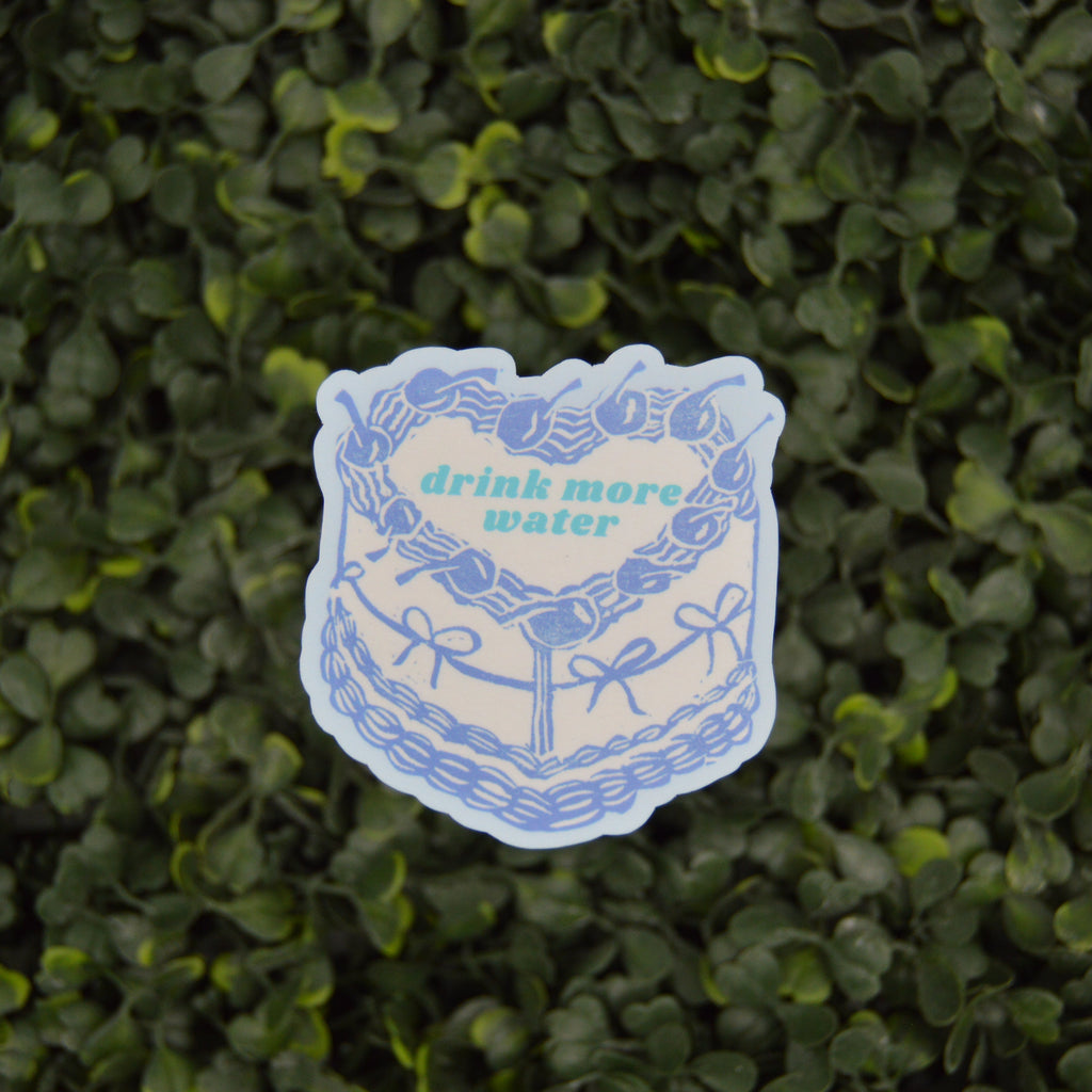 A die-cut sticker featuring a blue line art illustration of a cake with the text 'drink more water' in green, placed on a background of green foliage.