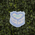 A die-cut sticker featuring a blue line art illustration of a cake with the text 'drink more water' in green, placed on a background of green foliage.