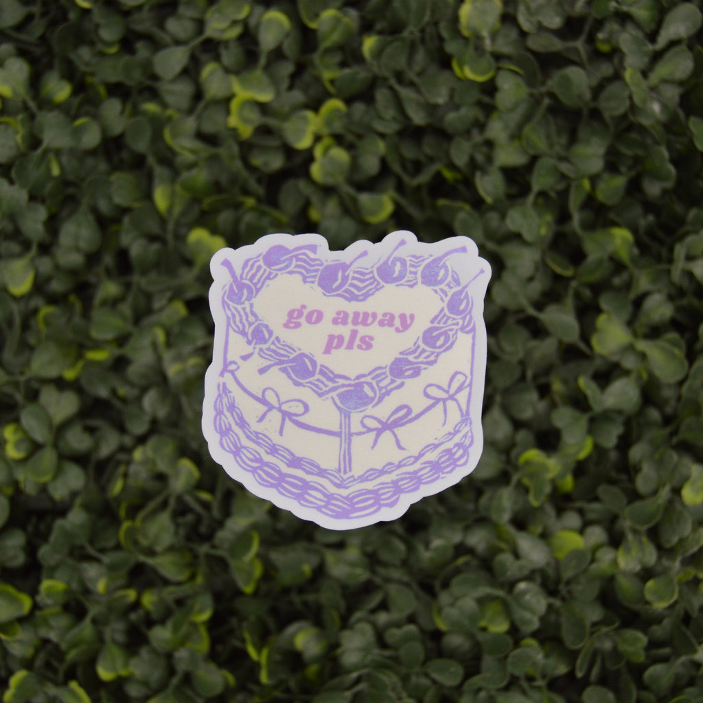 A die-cut sticker featuring a purple line art illustration of a cake with a heart-shaped center that reads 'go away pis' in pink text, shown against a background of green leafy hedge.