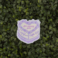A die-cut sticker featuring a purple line art illustration of a cake with a heart-shaped center that reads 'go away pis' in pink text, shown against a background of green leafy hedge.