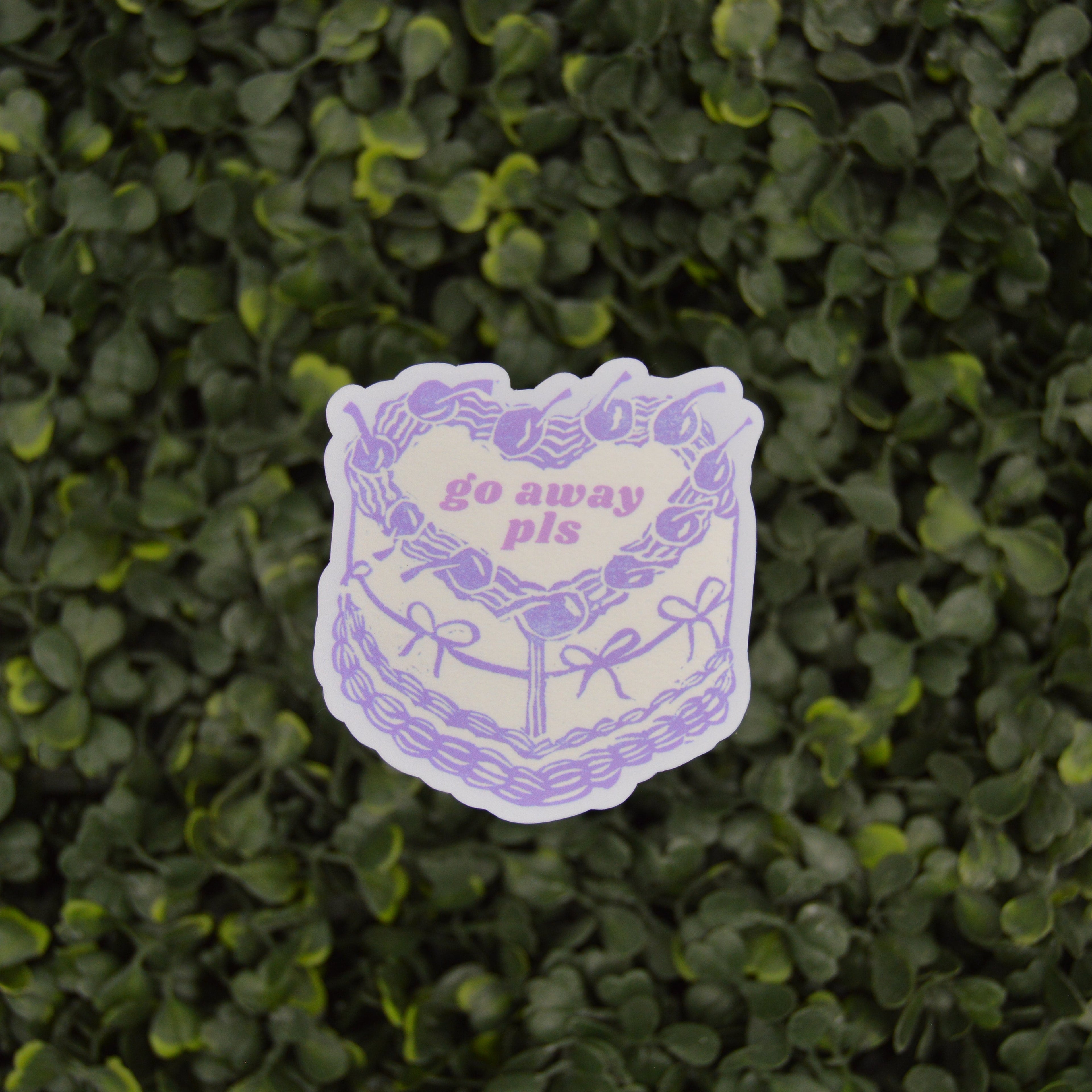 A die-cut sticker featuring a purple line art illustration of a cake with a heart-shaped center that reads 'go away pis' in pink text, shown against a background of green leafy hedge.