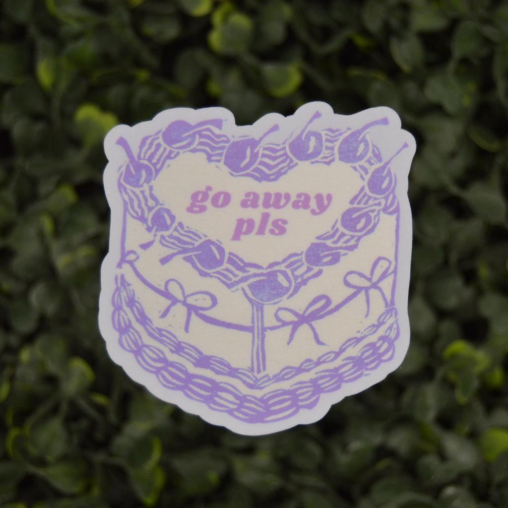 A die-cut sticker featuring a purple line-art illustration of a cake with the text 'go away pls' in purple, shown against a blurred green leaf background.