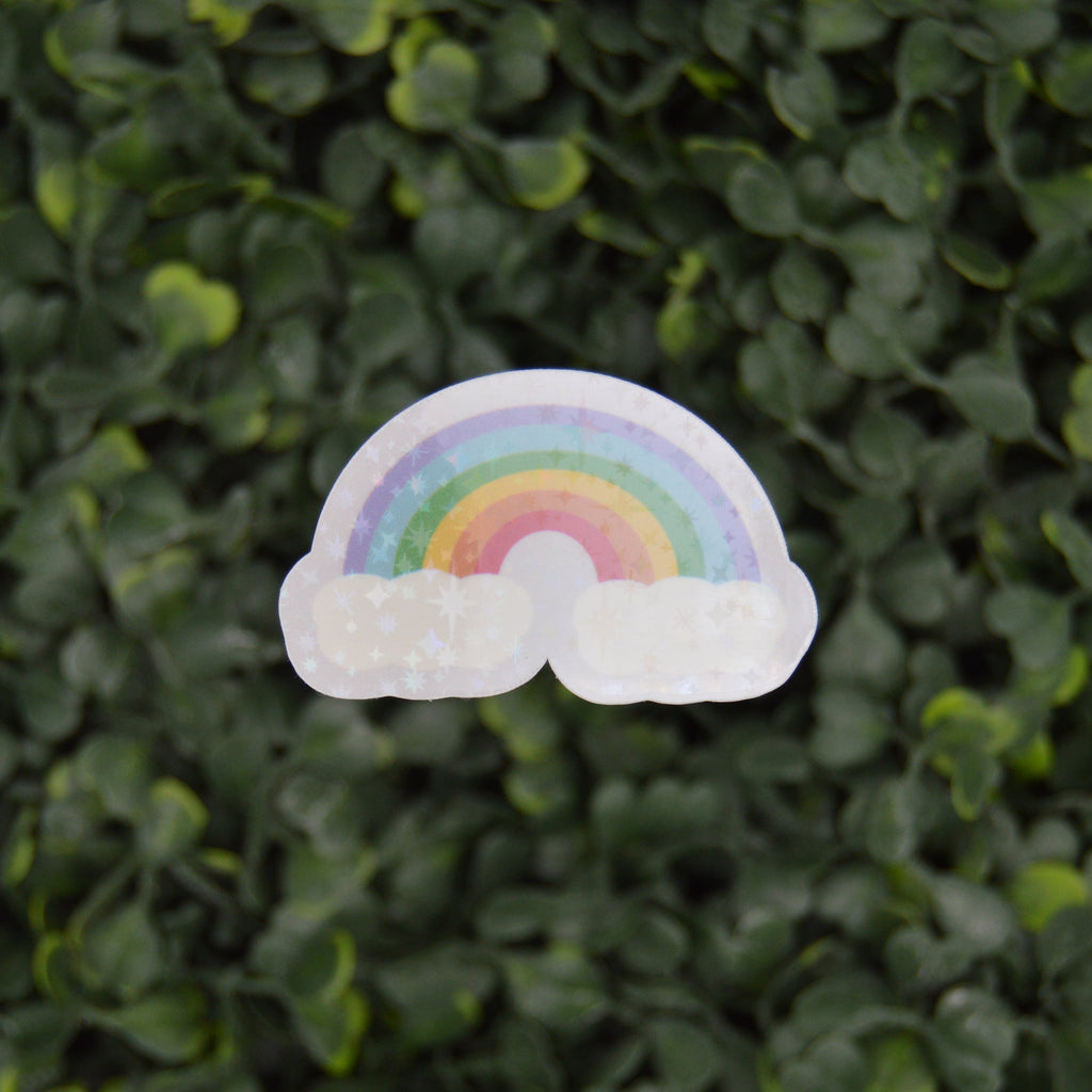 A small rainbow sticker with white clouds on either side, placed against a dense green leaf background.