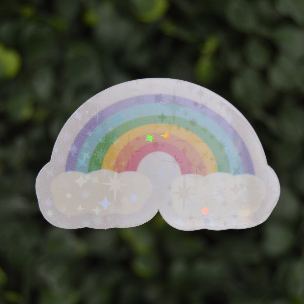 A die-cut sticker featuring a pastel rainbow and white clouds with a subtle holographic glitter finish, shown against a blurred green leaf background.