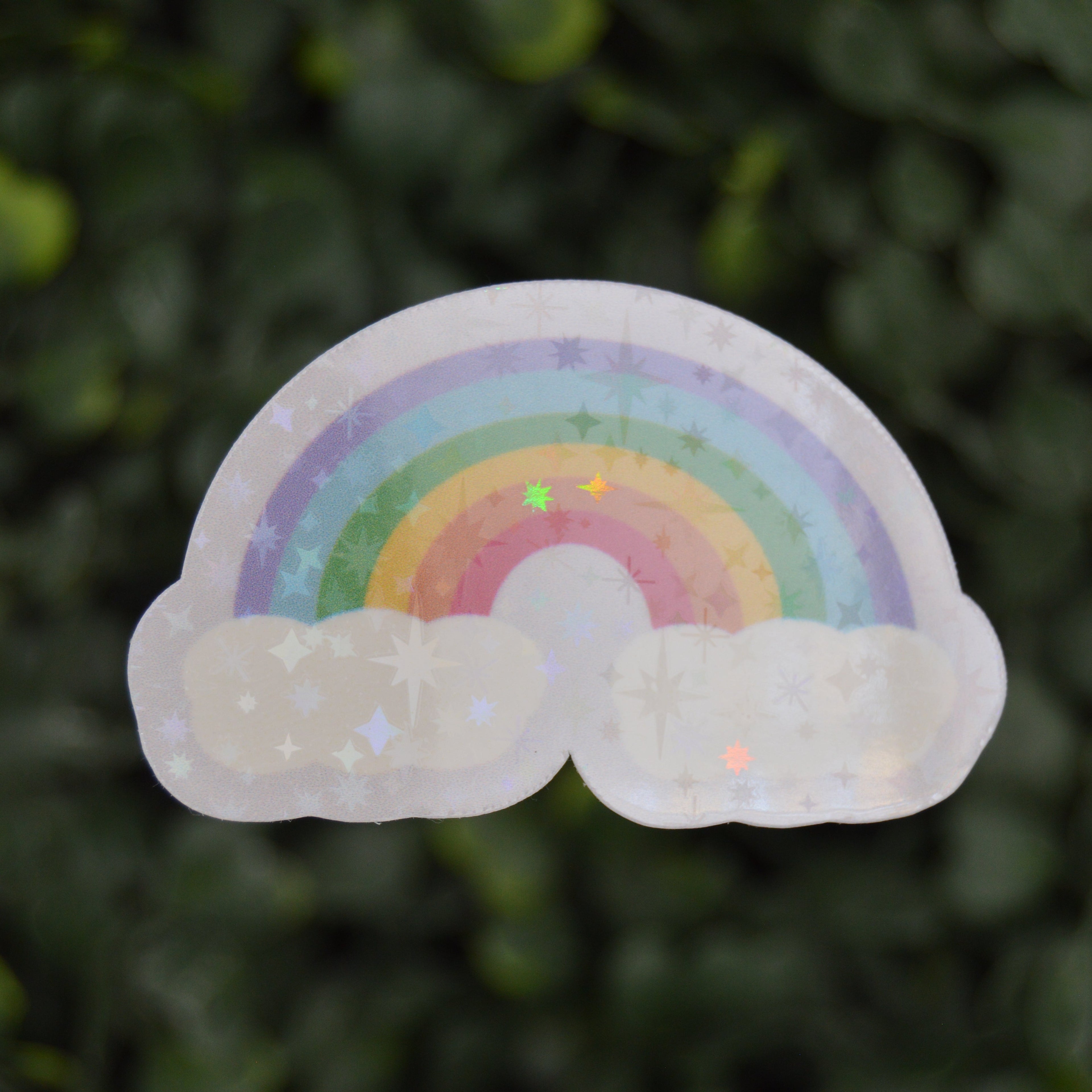 A die-cut sticker featuring a pastel rainbow and white clouds with a subtle holographic glitter finish, shown against a blurred green leaf background.