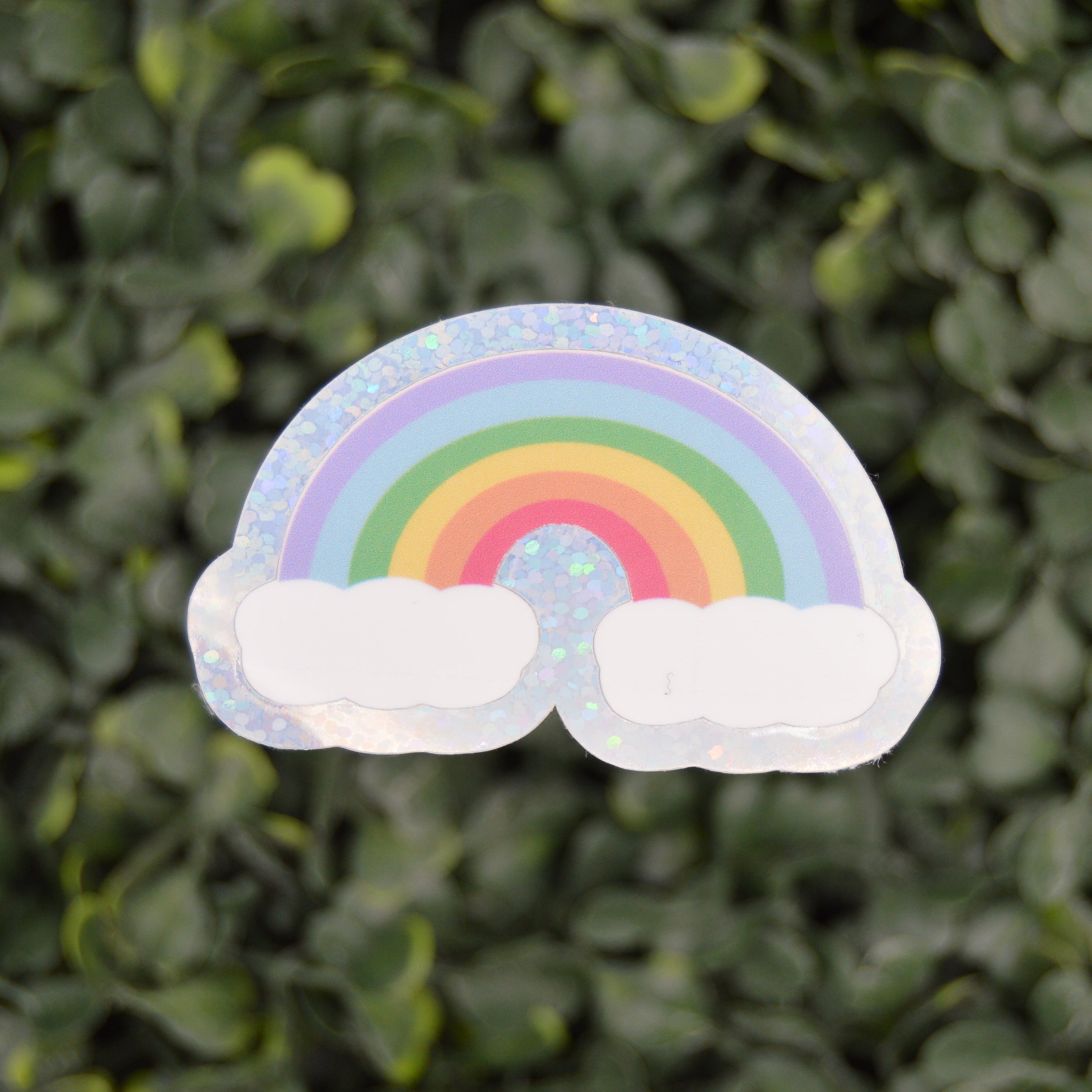A small rainbow sticker with two white clouds, placed against a blurred background of green leaves.