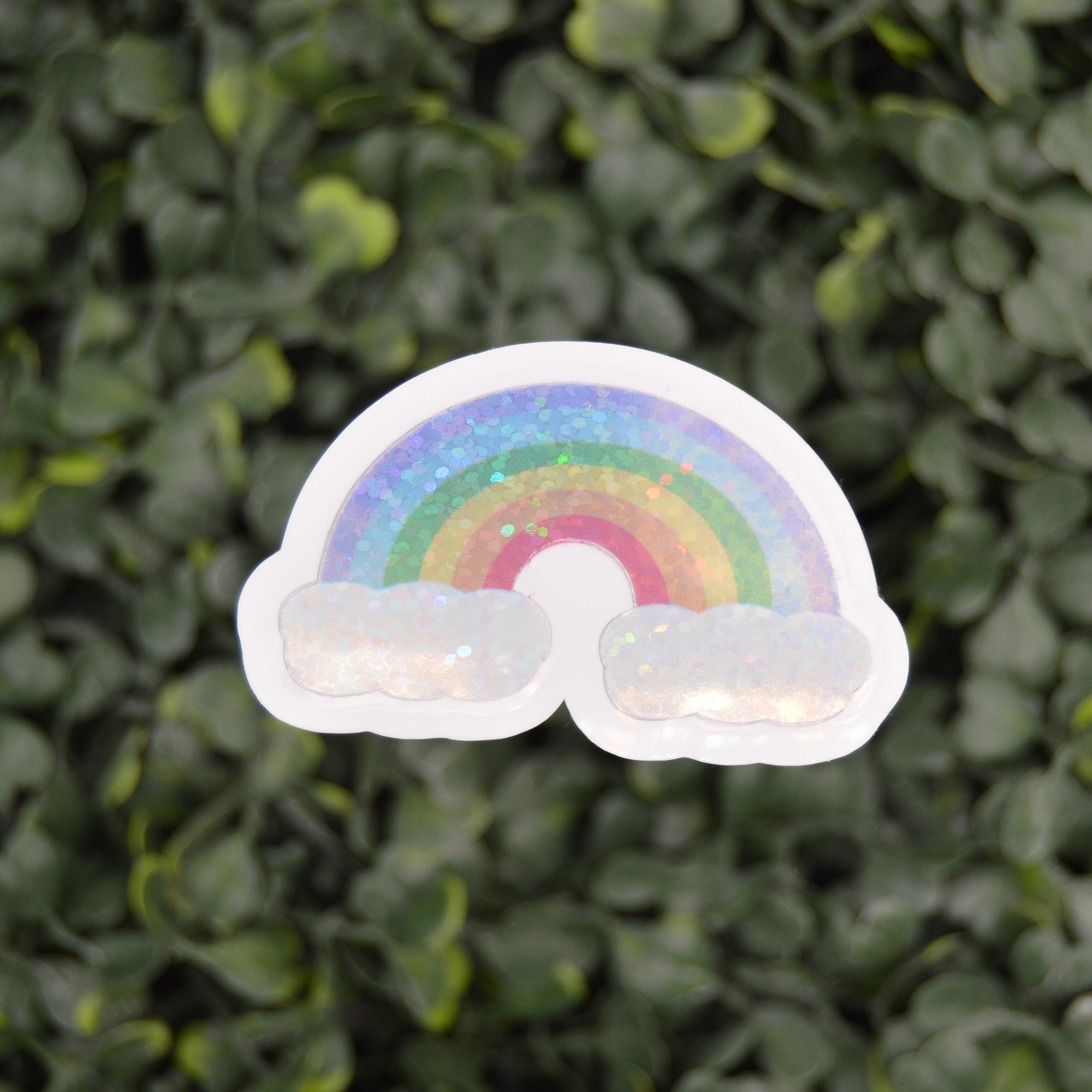 A die-cut sticker featuring a pastel rainbow arch above two fluffy white clouds, shown against a blurred green leaf background.