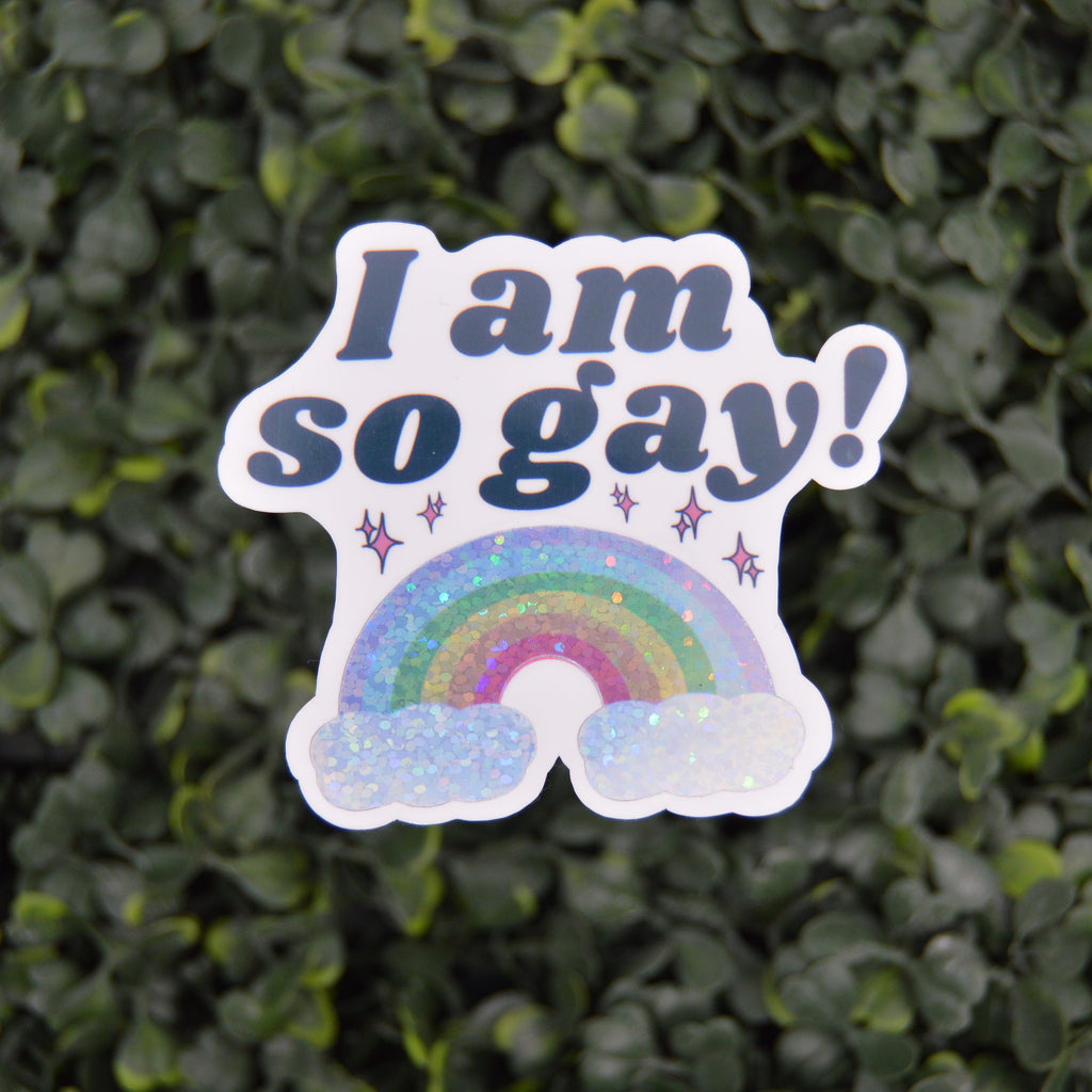 A die-cut sticker with the text 'I am so gay!' in dark blue lettering above a glittery rainbow and clouds, shown against a blurred green leaf background.