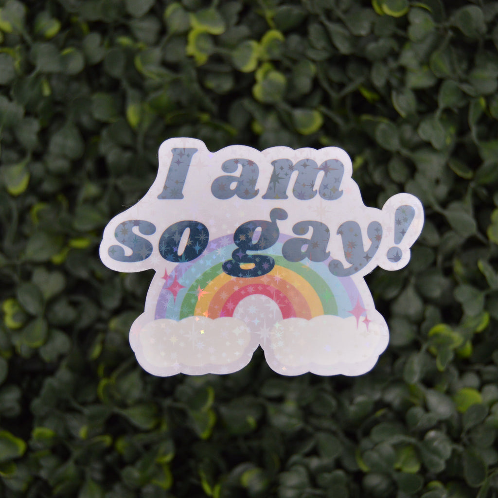 A die-cut sticker with the text 'I am so gay!' in glittery blue letters above a rainbow and clouds, shown against a blurred green leaf background.