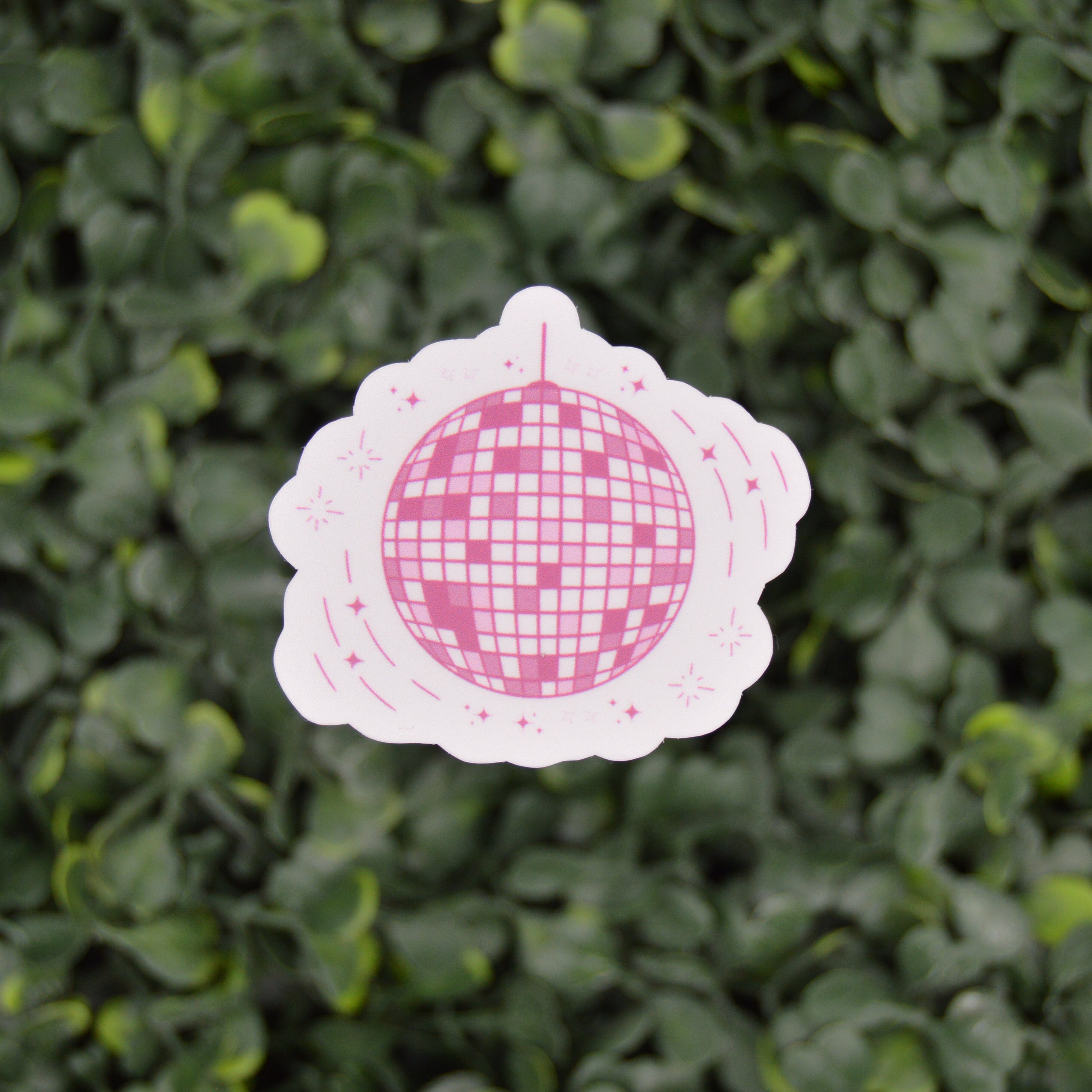 A die-cut sticker featuring a pink and white disco ball design on a scalloped white border, shown against a blurred green leafy background.