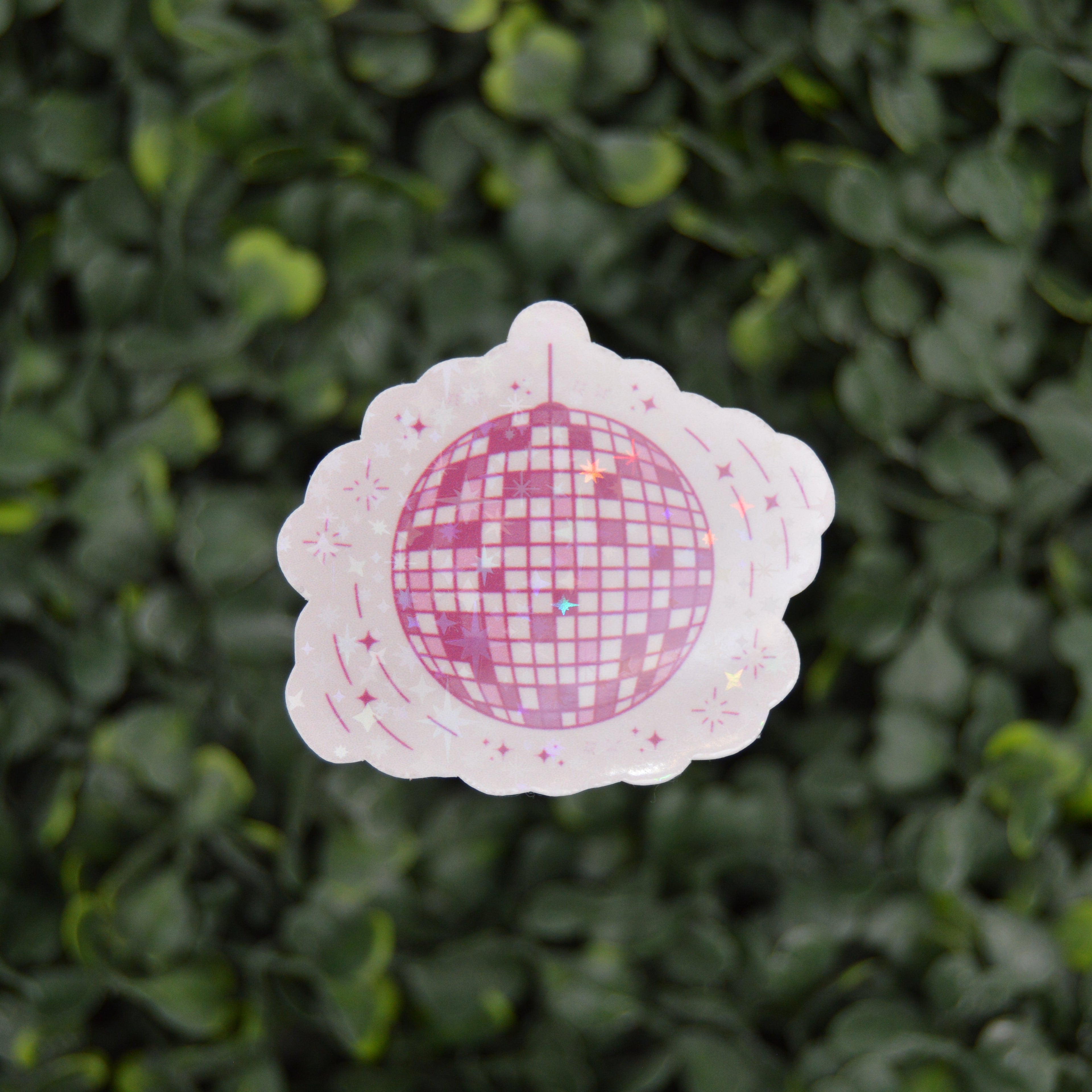 A scalloped white sticker or decal featuring a pink and white disco ball design, shown against a blurred green leaf background.