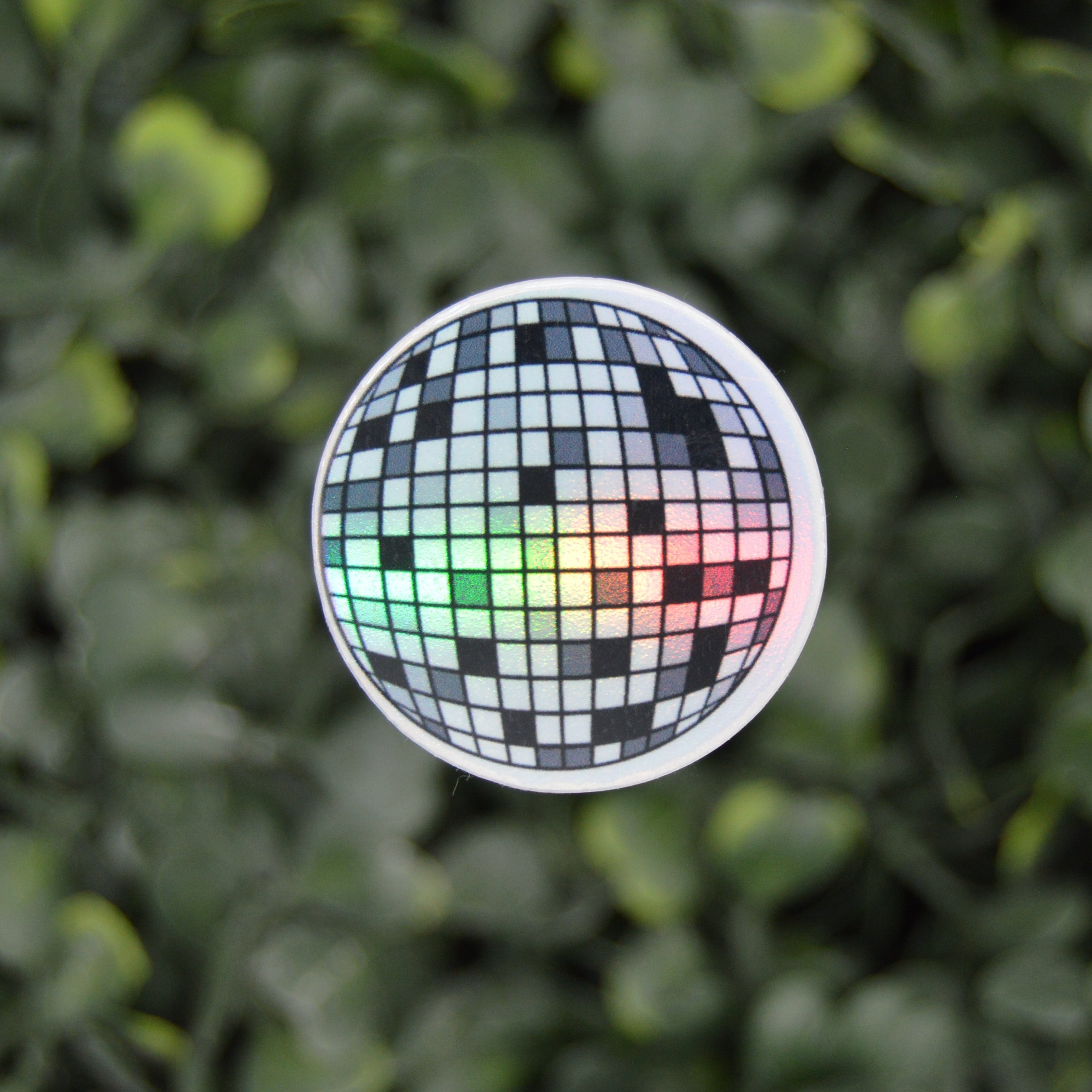 A round sticker featuring a black and white disco ball design with a green, yellow, and red light effect, shown against a blurred green leaf background.