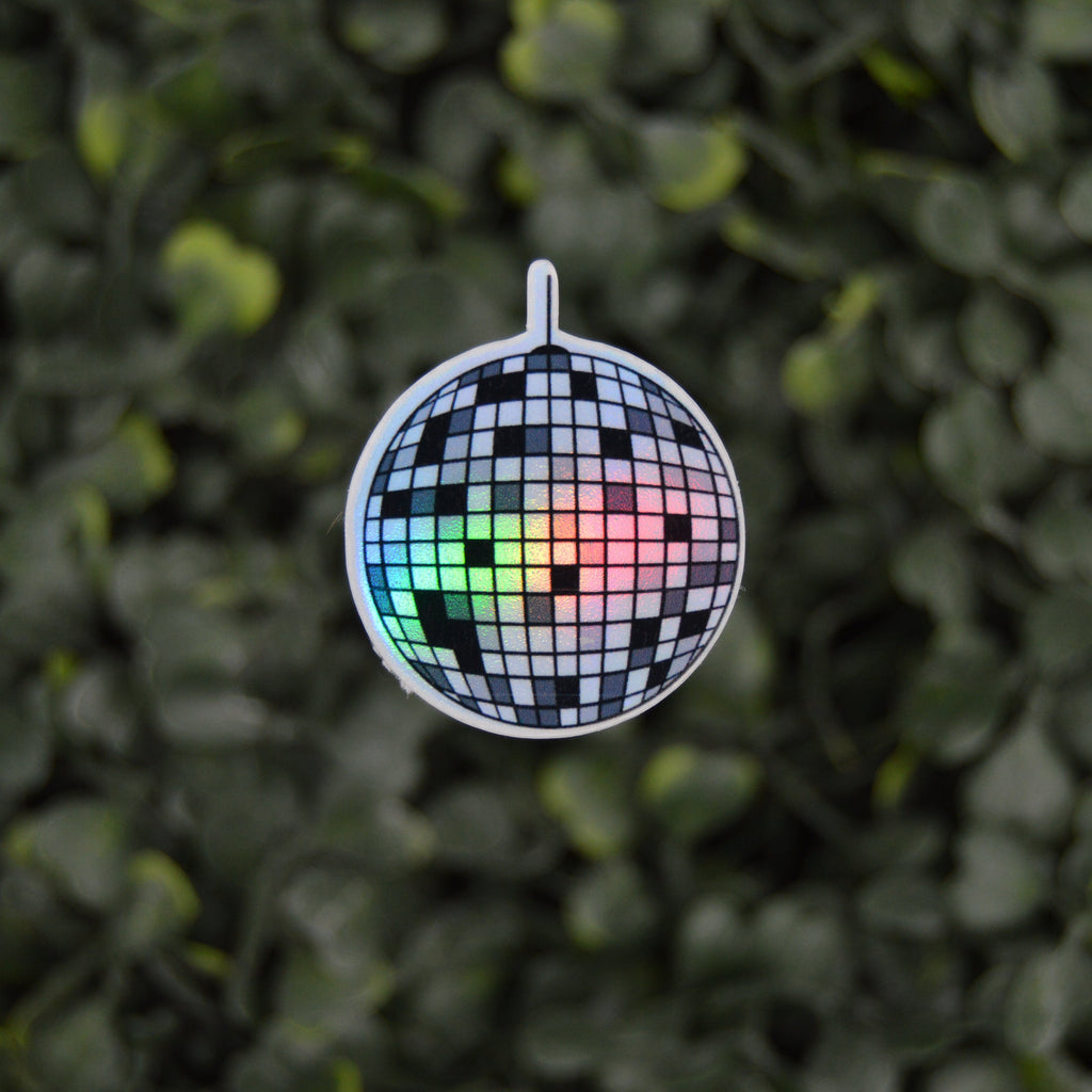 A mini disco ball charm with a black and white checkered mirror tile pattern and a small rainbow light reflecting through the center, hanging against a blurred green leaf background.