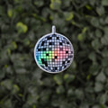 A mini disco ball charm with a black and white checkered mirror tile pattern and a small rainbow light reflecting through the center, hanging against a blurred green leaf background.