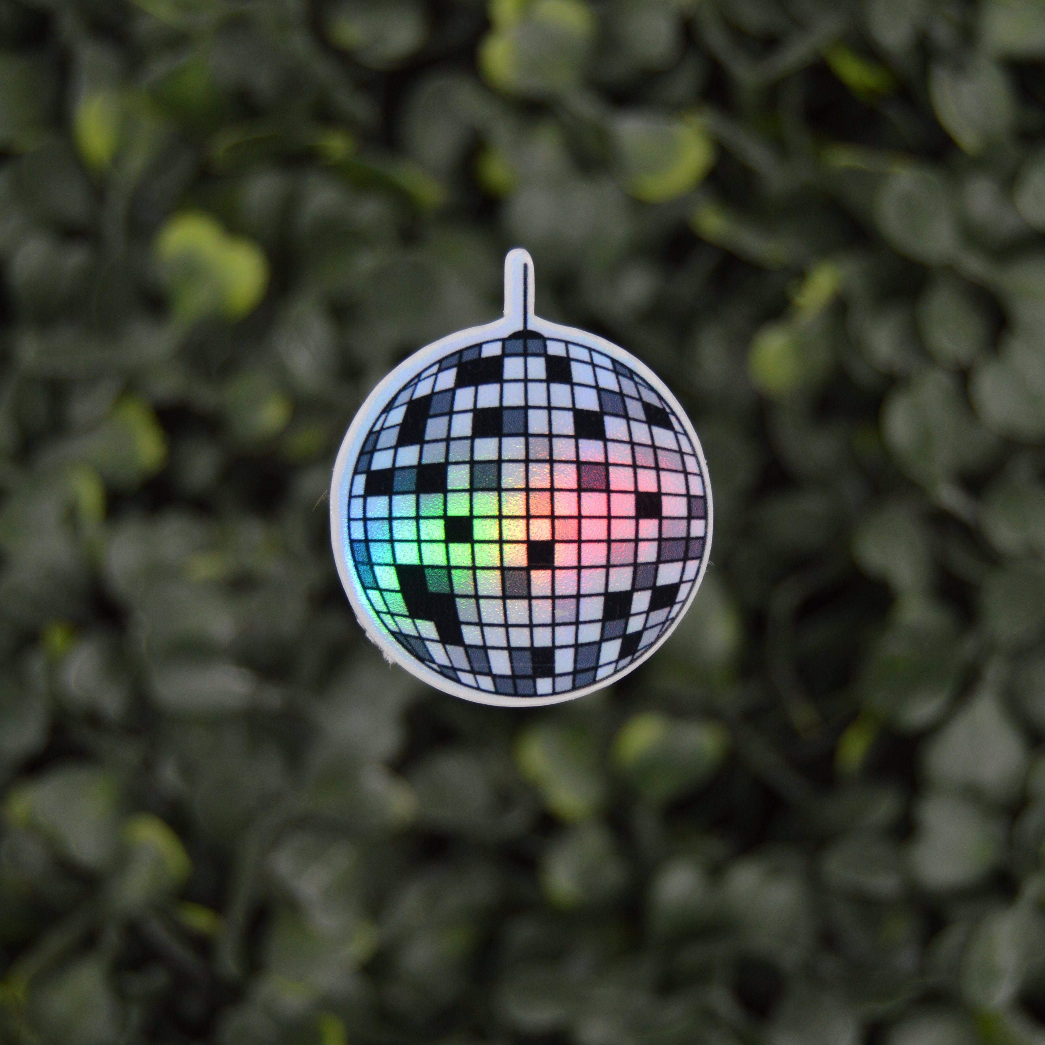 A mini disco ball charm with a black and white checkered mirror tile pattern and a small rainbow light reflecting through the center, hanging against a blurred green leaf background.