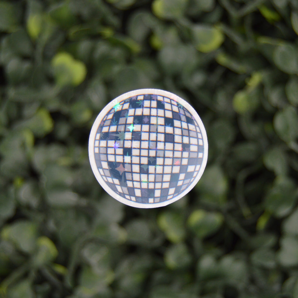 A round disco ball sticker hanging against a dense green leaf wall background.