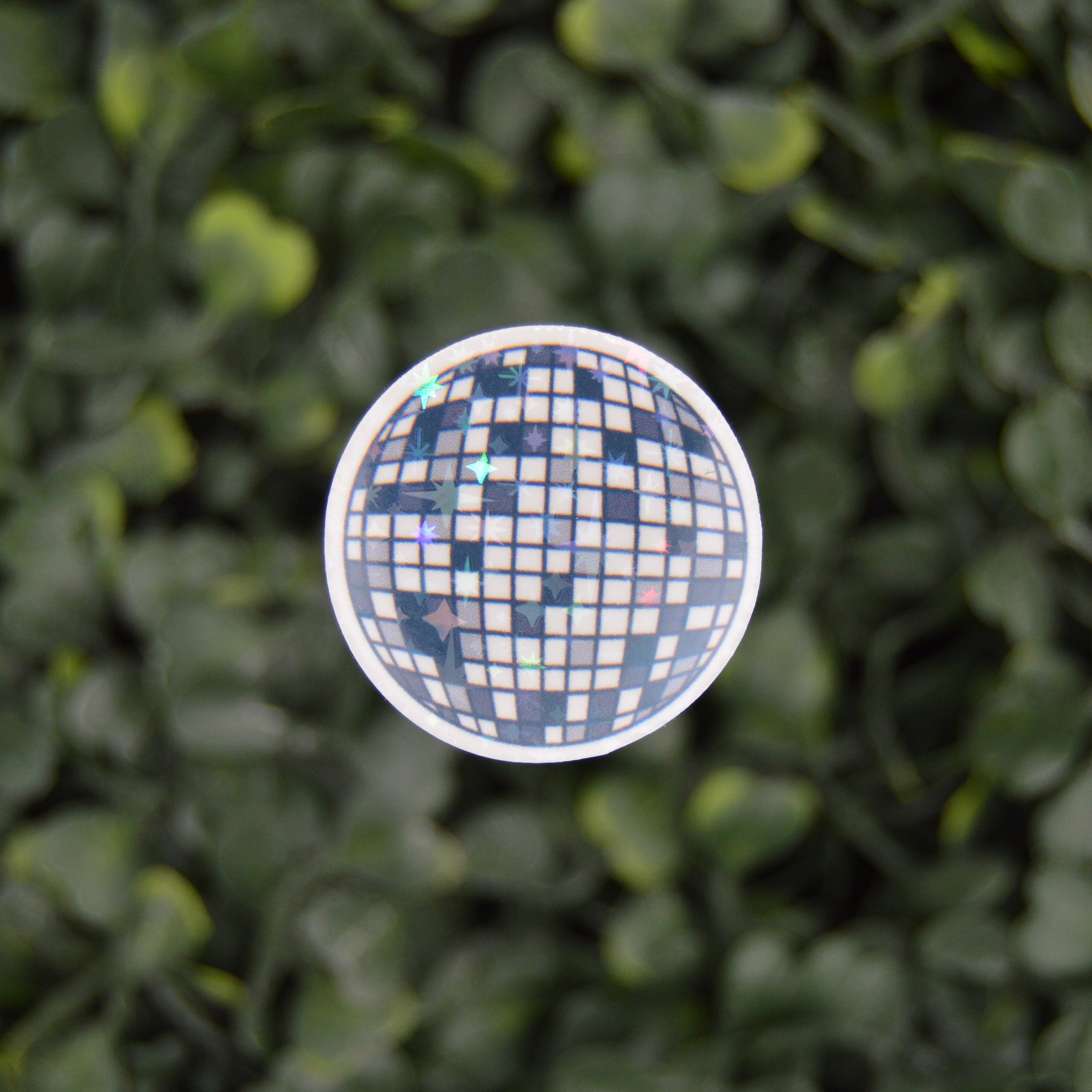 A round disco ball sticker hanging against a dense green leaf wall background.