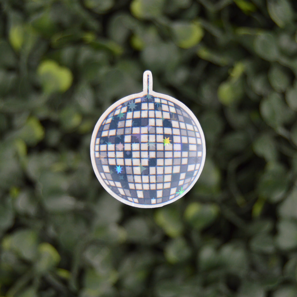 A round disco ball pendant with a blue and white checkered pattern and scattered blue and yellow glitter, suspended against a blurred green leaf background.