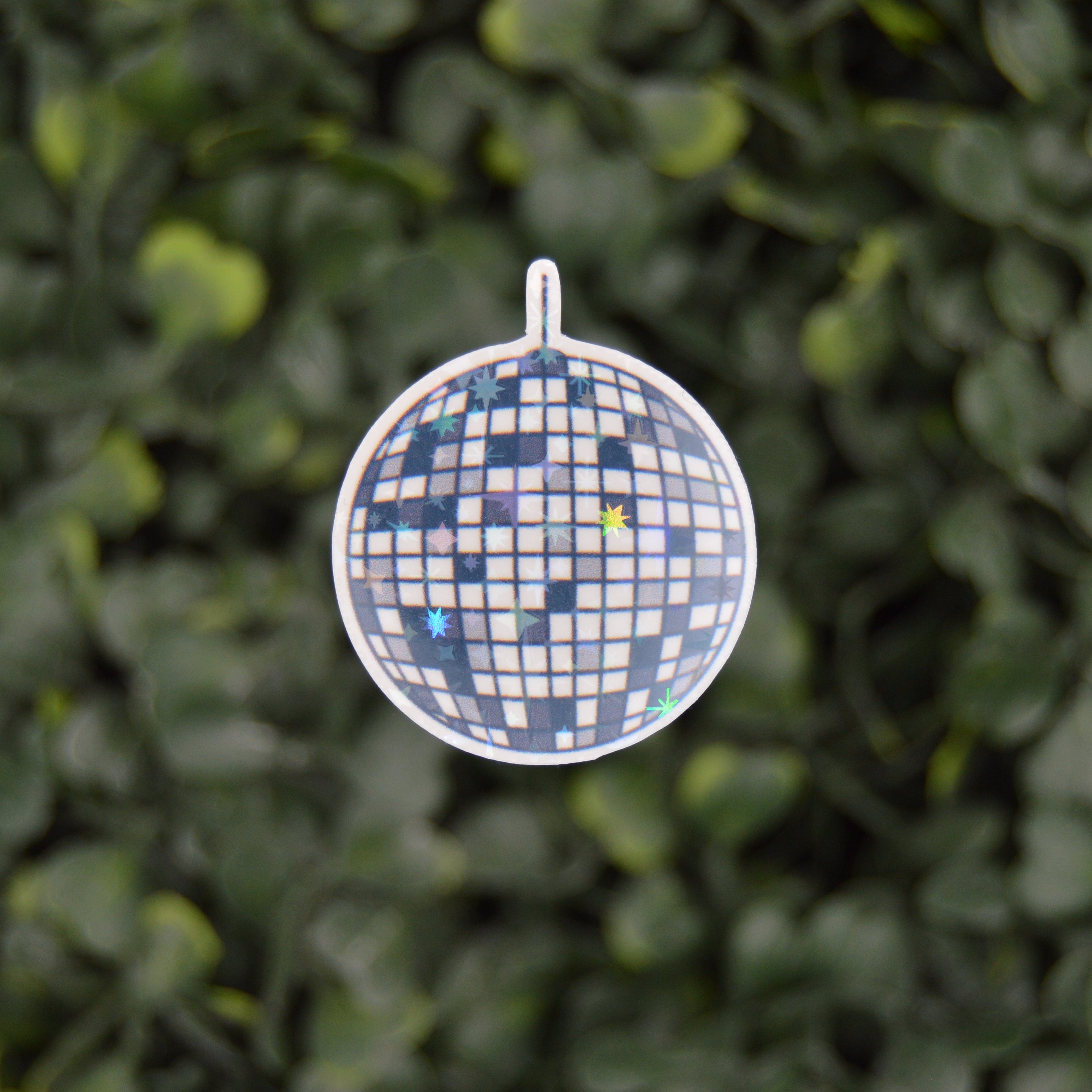 A round disco ball pendant with a blue and white checkered pattern and scattered blue and yellow glitter, suspended against a blurred green leaf background.