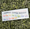 A rectangular white bumper sticker with the text 'I SUPPORT ZIPPER MERGES!' and cartoon cars, placed on a bed of green leaves.