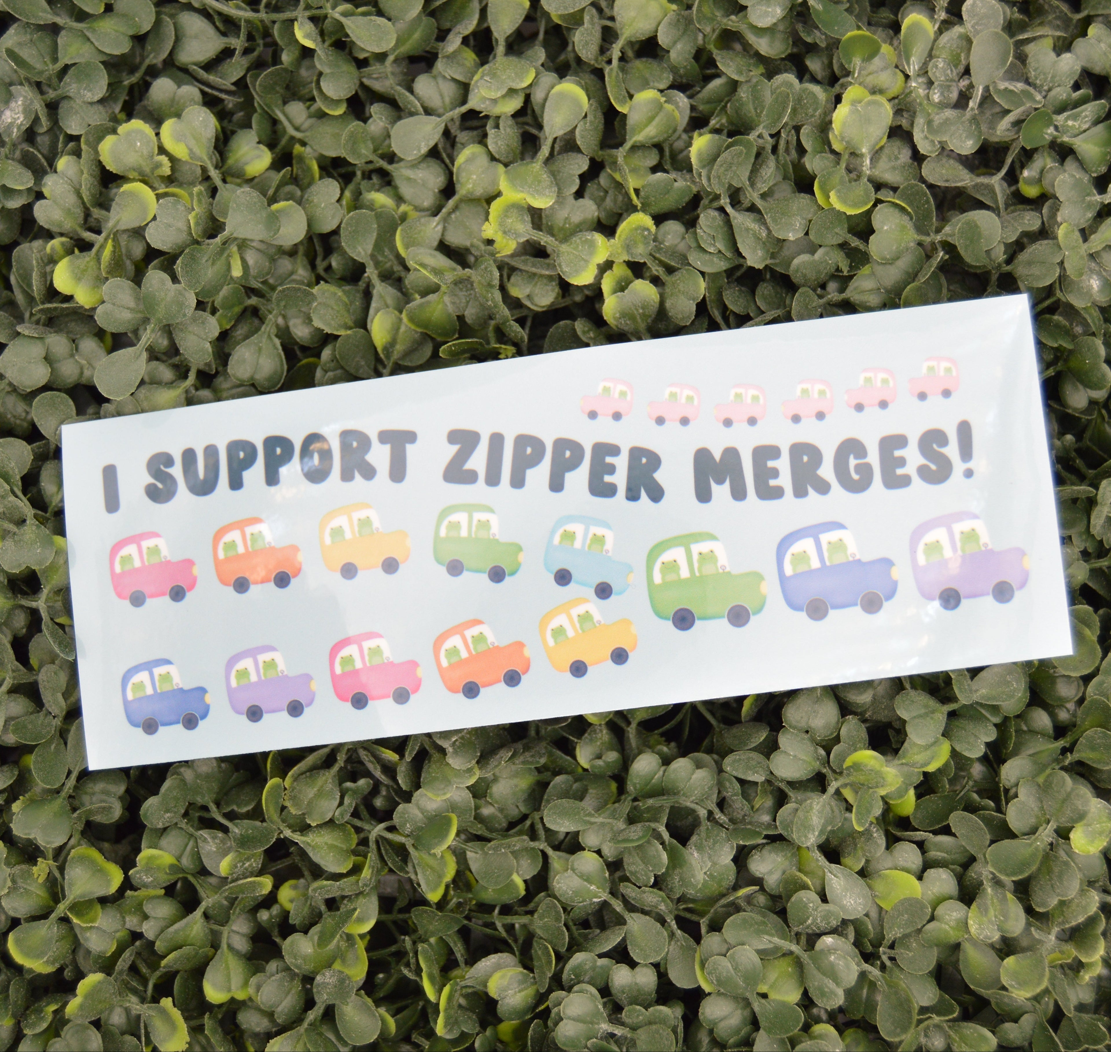 A rectangular white bumper sticker with the text 'I SUPPORT ZIPPER MERGES!' and cartoon cars, placed on a bed of green leaves.