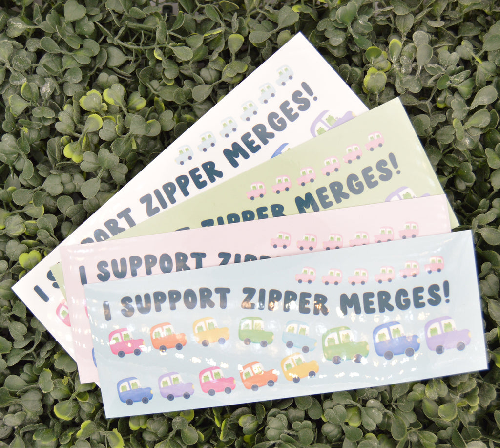 Three rectangular bumper stickers with the text 'I SUPPORT ZIPPER MERGES!' in dark blue letters, decorated with rows of small cartoon cars in various colors. The stickers are fanned out on a bed of green leaves.